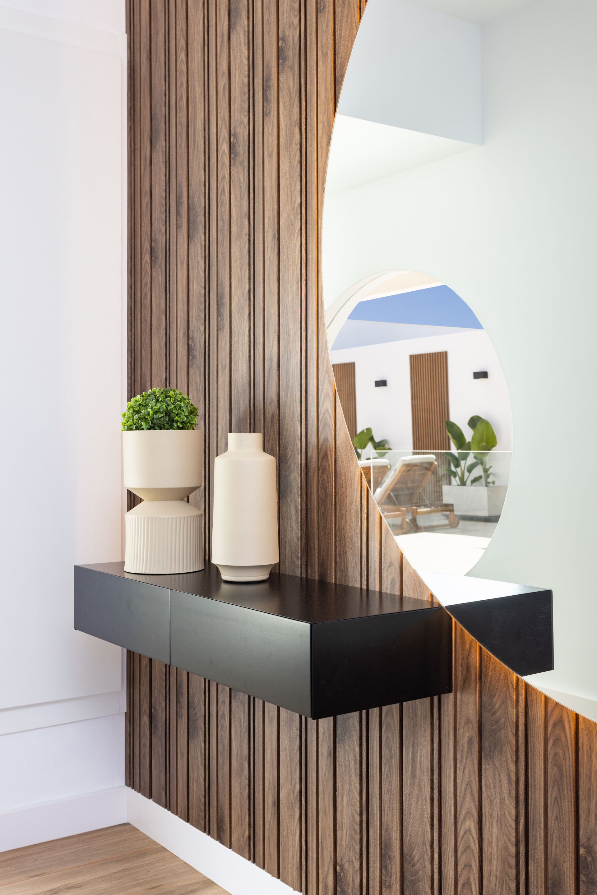 Entrada moderna con listones de madera verticales, un estante negro y jarrones decorativos sobre una pared blanca.