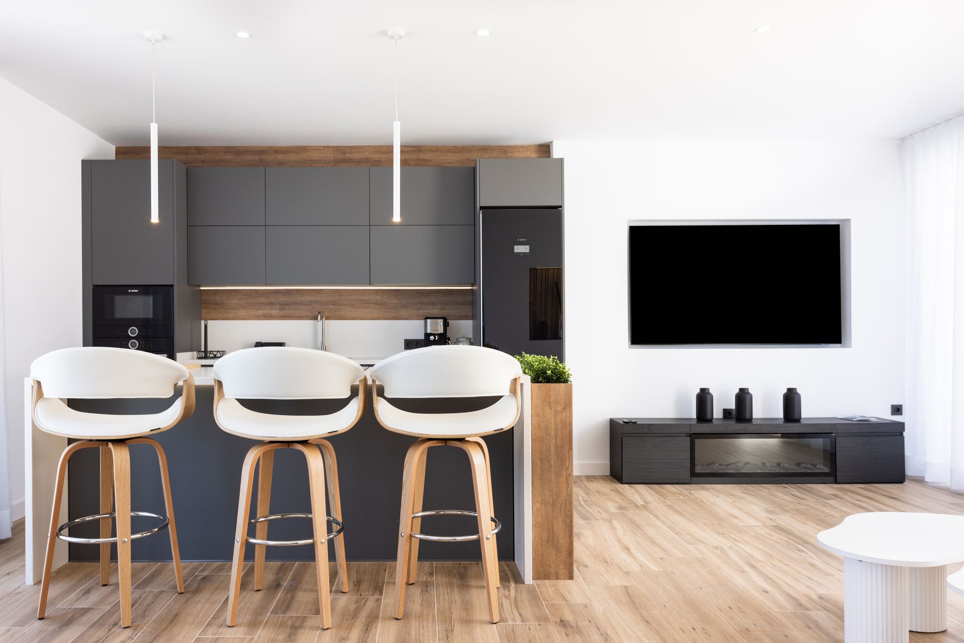 Modern kitchen and living room with white bar stools, wood floors, and wall-mounted TV