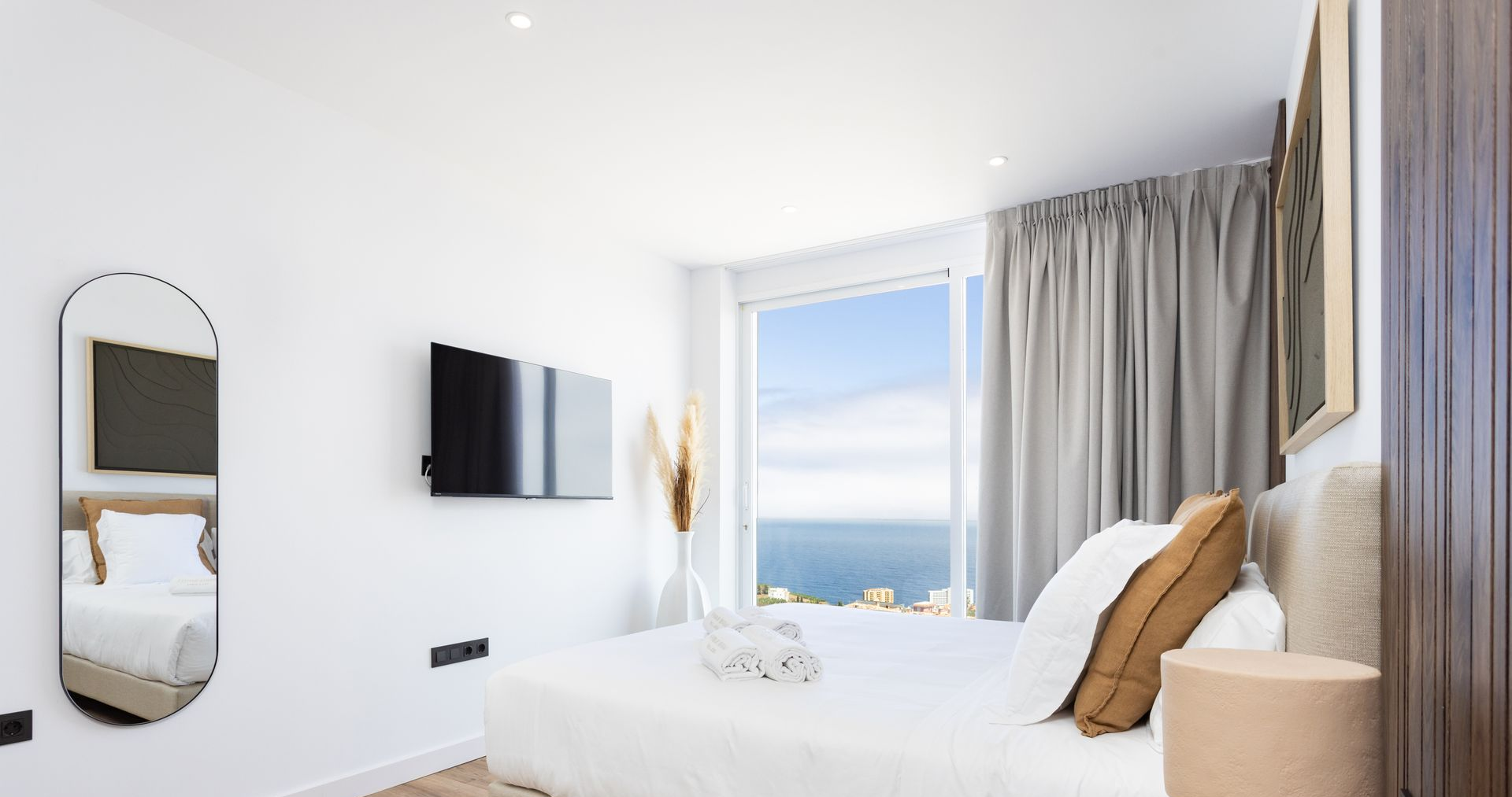 Bright minimalist bedroom with a bed, wall-mounted TV, mirror, and window overlooking the sea