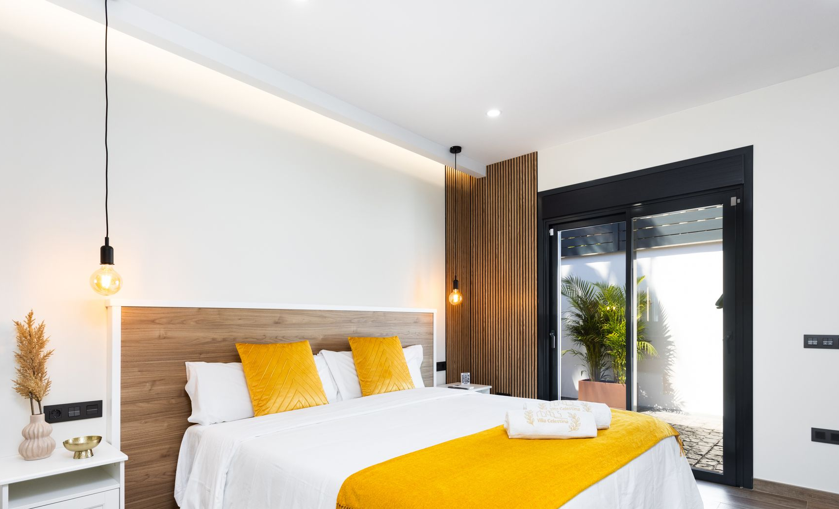Modern bedroom with white bed, mustard accents, wood headboard, and balcony doors opening to greenery