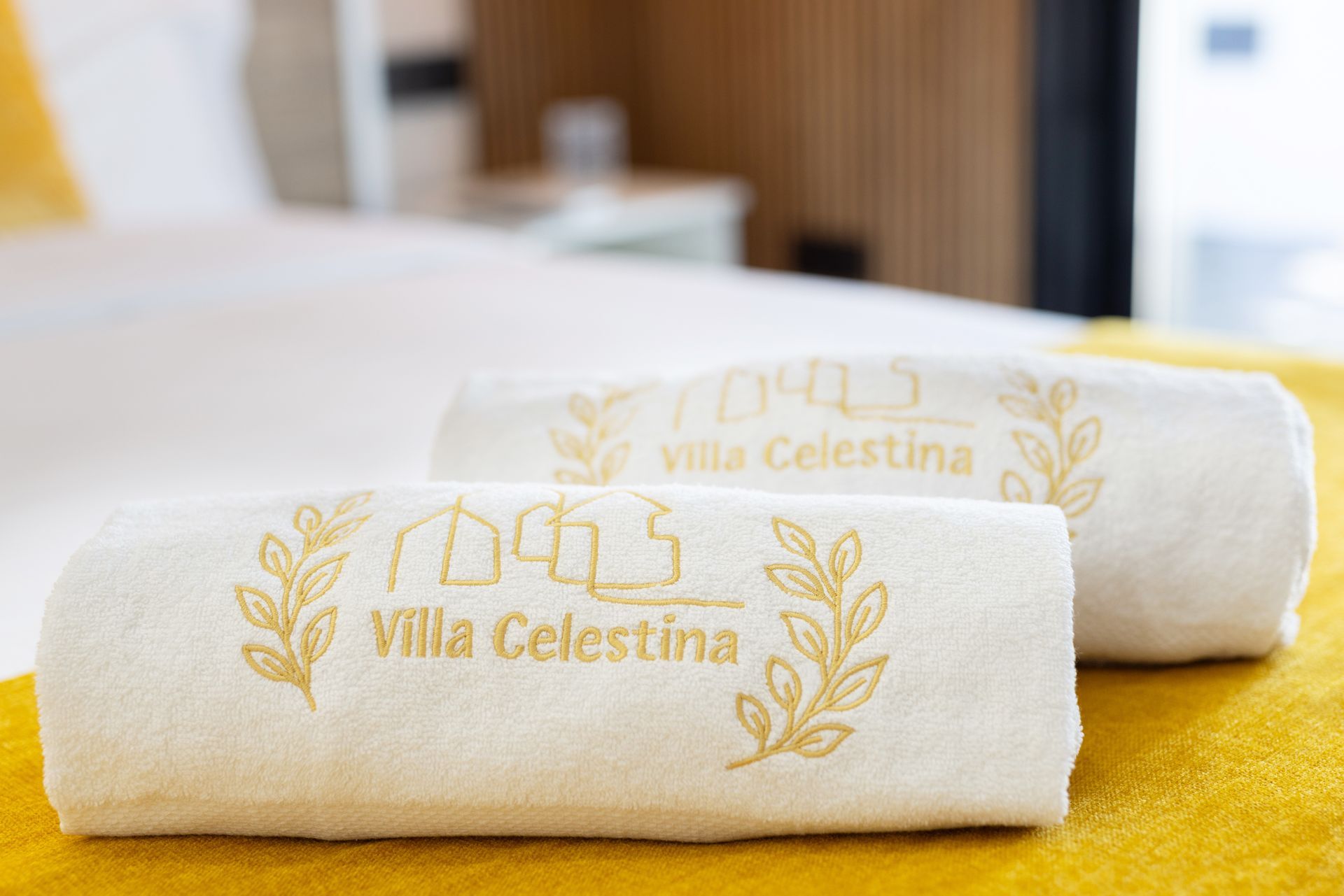 White rolled towels with gold “Villa Celestina” embroidery on a bed in a hotel room