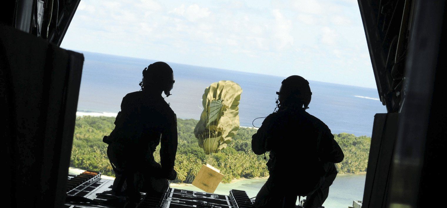 Two soldiers kneeling at the back door of a flying helicopter dropping a package with a parachute