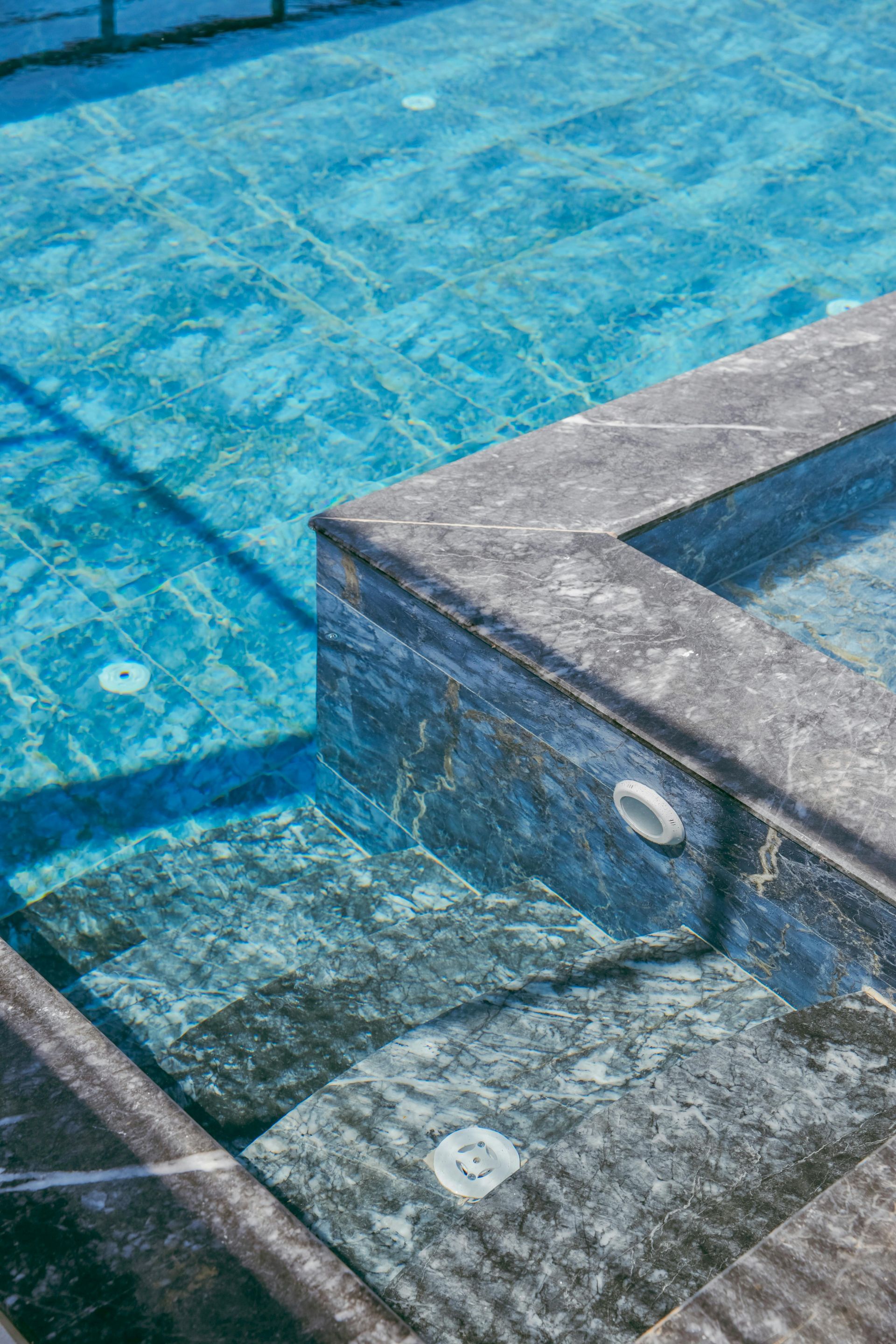 Stone steps descending into a blue tiled swimming pool, with clear water and sunlit shadows
