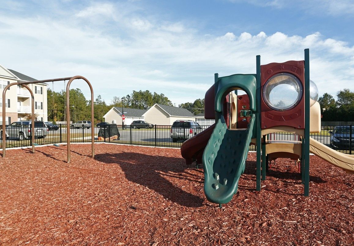 Playground with slides and climbing structure on mulch, fenced area near apartment buildings at Jack Britt.