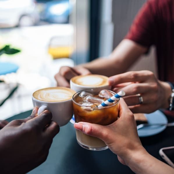 Three hands toast with two latte mugs and a glass of iced coffee in a bright cafe setting.
