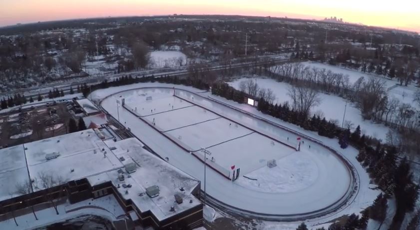 Aerial view of a snow-covered outdoor skating rink with surrounding trees and buildings under a dusky sky.