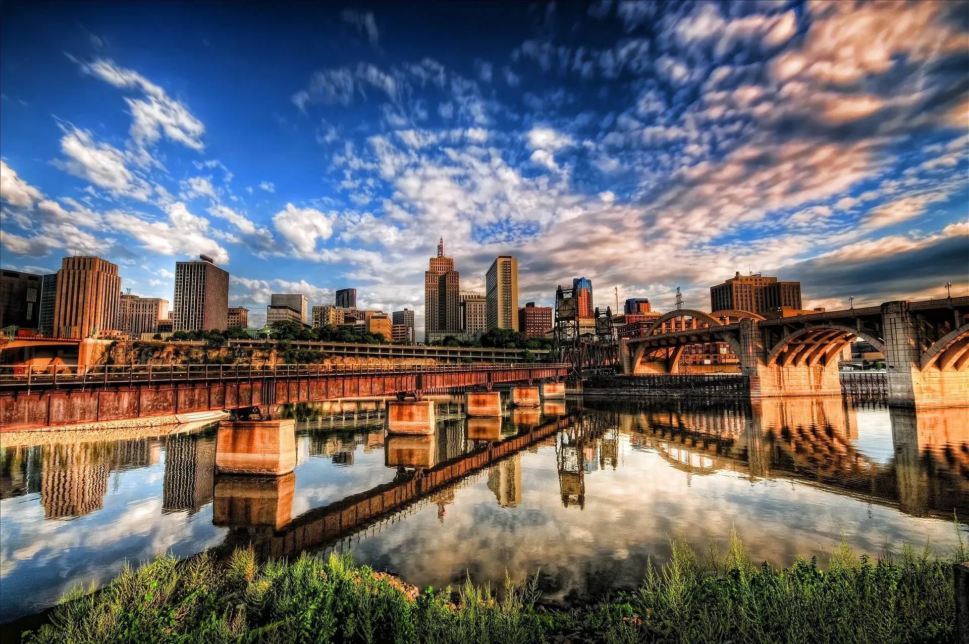 City skyline reflected in water, with bridge, trees, and dramatic cloudy sky.