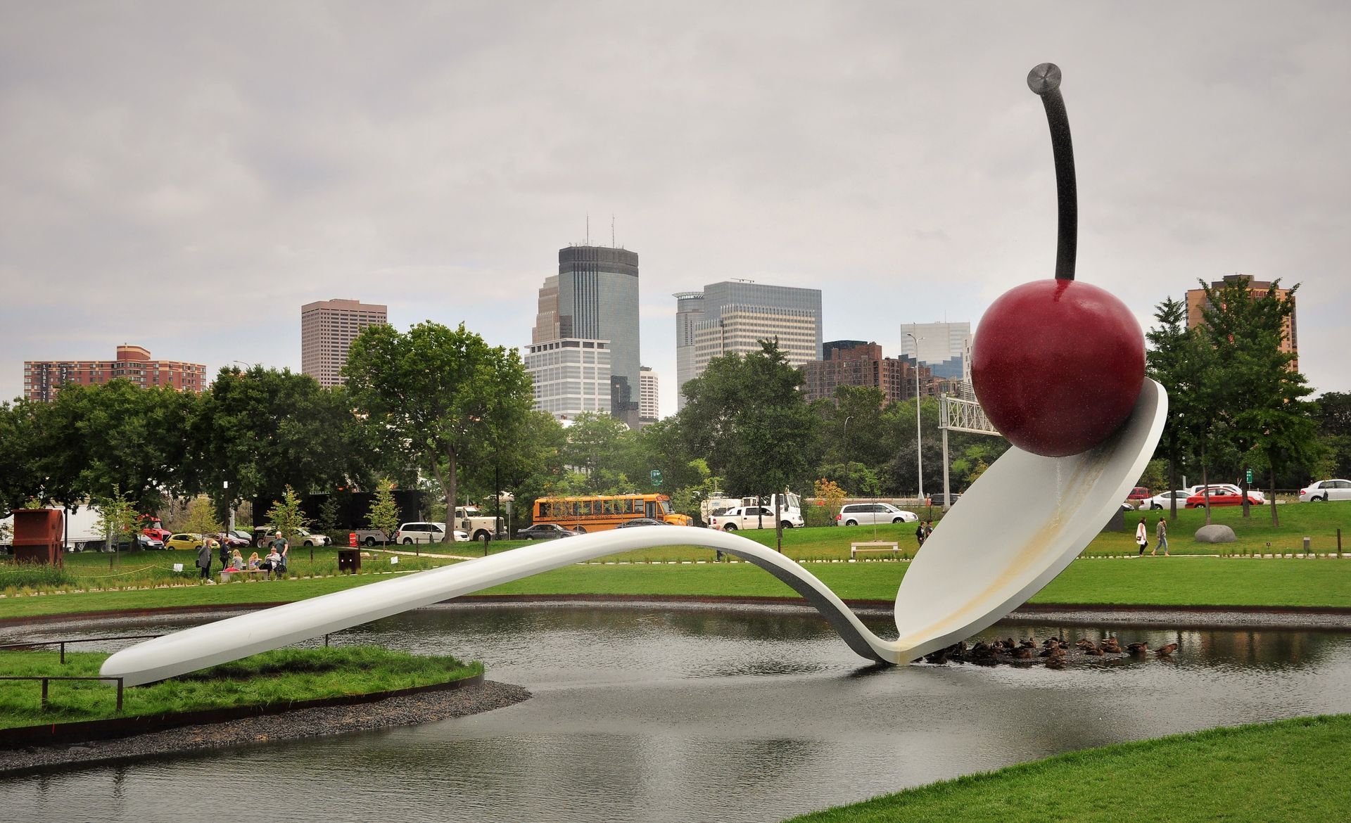 Giant white spoon and red cherry sculpture in a pond, with a city skyline backdrop.