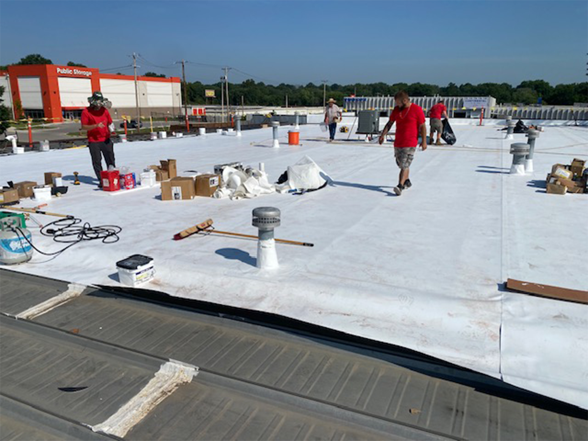 Workers on a white commercial roof. One welds, others walk. Boxes and tools are scattered. Bright sunlight.