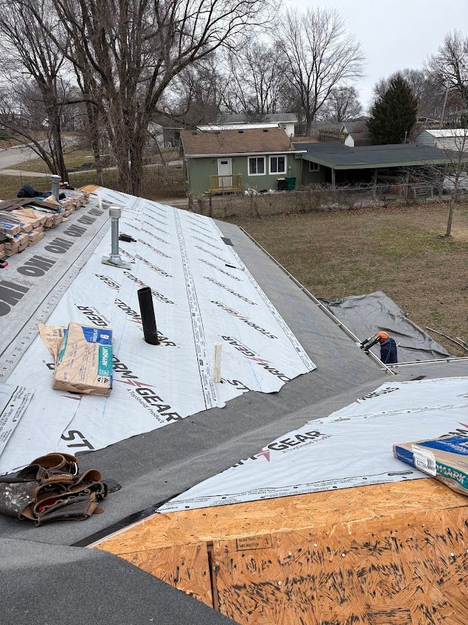 Roofer using a nail gun to install asphalt shingles on a roof under a bright blue sky.