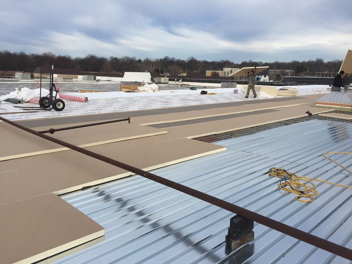 Two roofers installing black shingles on a roof under a bright blue sky.