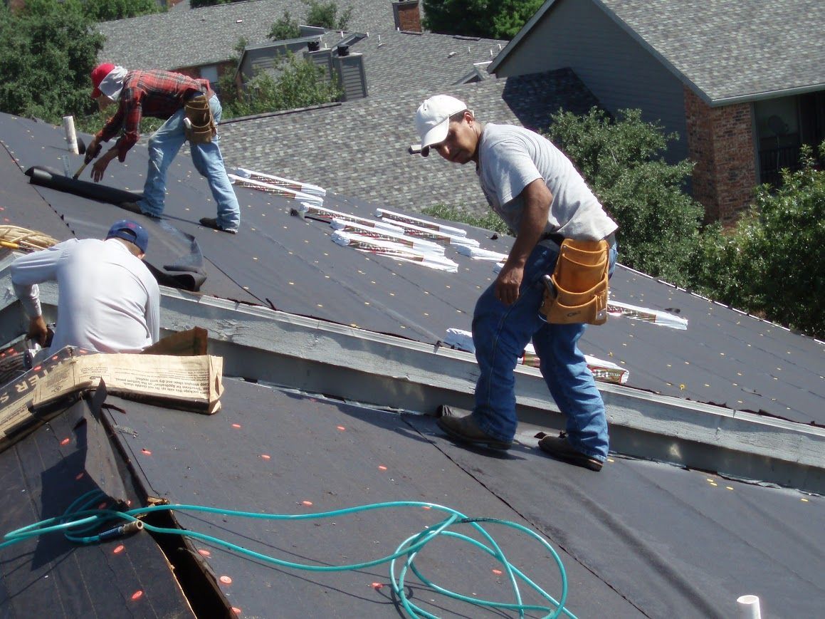 Two roofers installing black shingles on a roof under a bright blue sky.