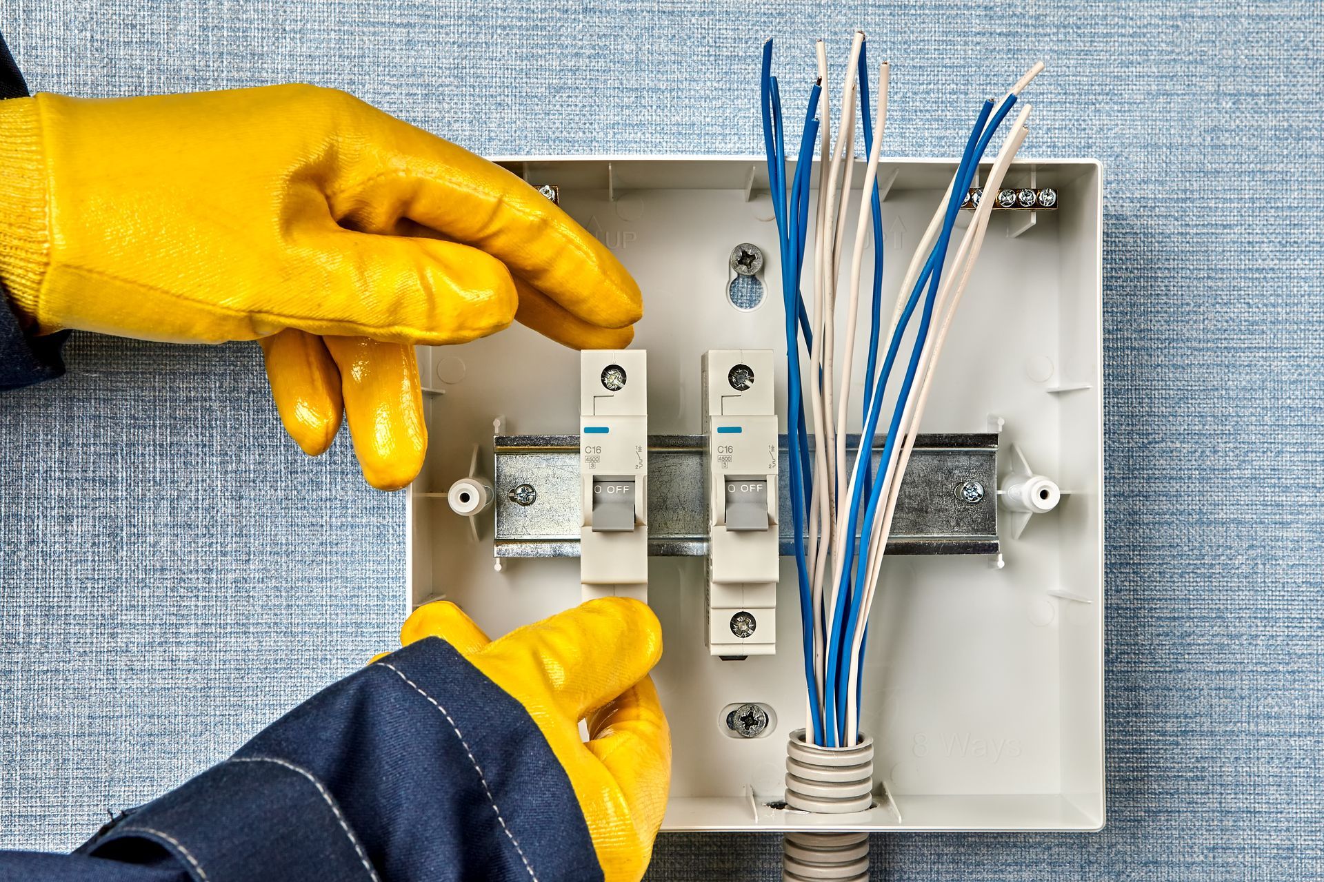 Hands in yellow gloves installing electrical circuit breakers in a white box on a blue wall.