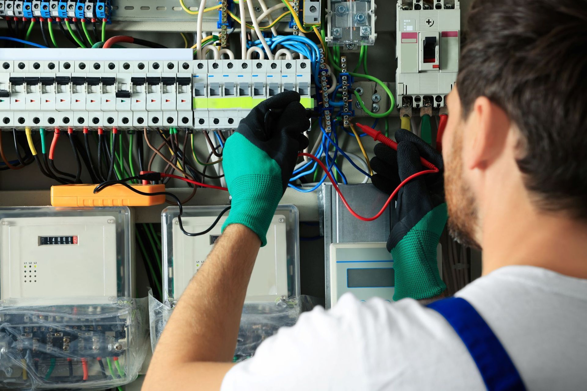 Electrician working on electrical panel, wearing gloves, using multimeter.