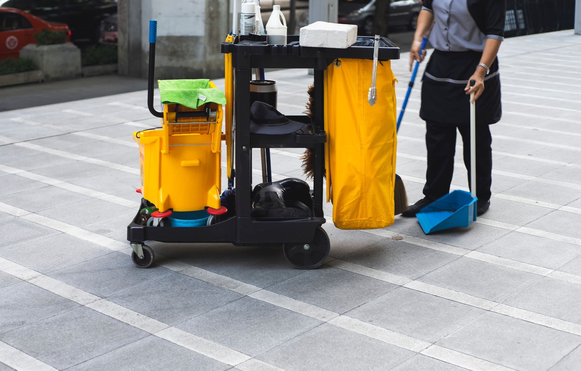 Janitor sweeping outdoor walkway next to cleaning cart. Yellow buckets, black cart, and blue dustpan.