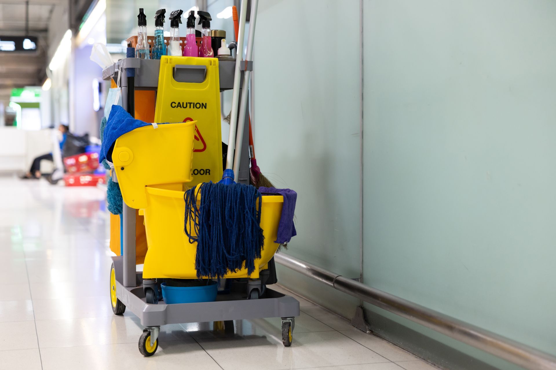 Fully stocked cleaning cart with mop, bucket, and supplies in a building corridor.