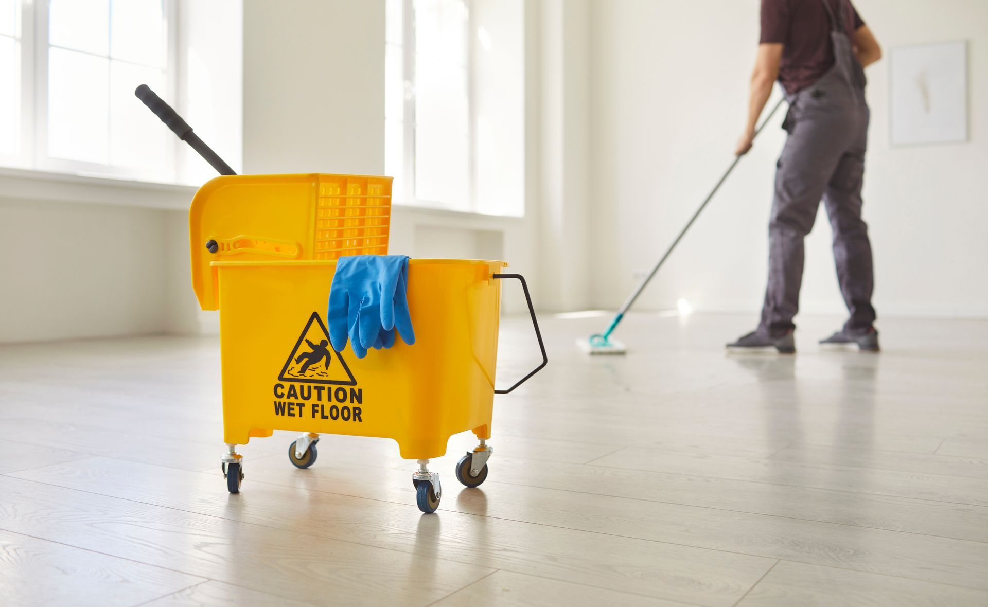 Janitor mopping a spacious room with a caution wet floor bucket placed nearby.