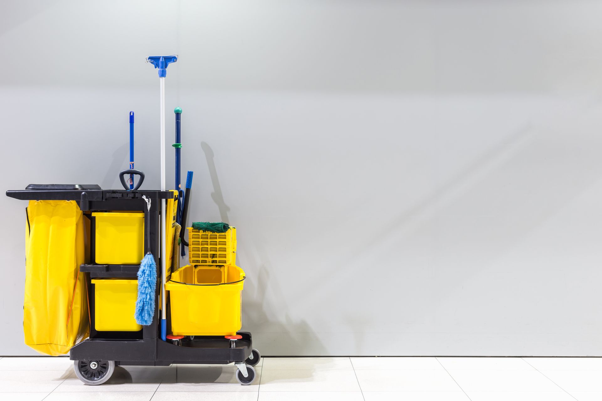 Yellow cleaning cart with mop, bucket, and supplies against a grey wall.