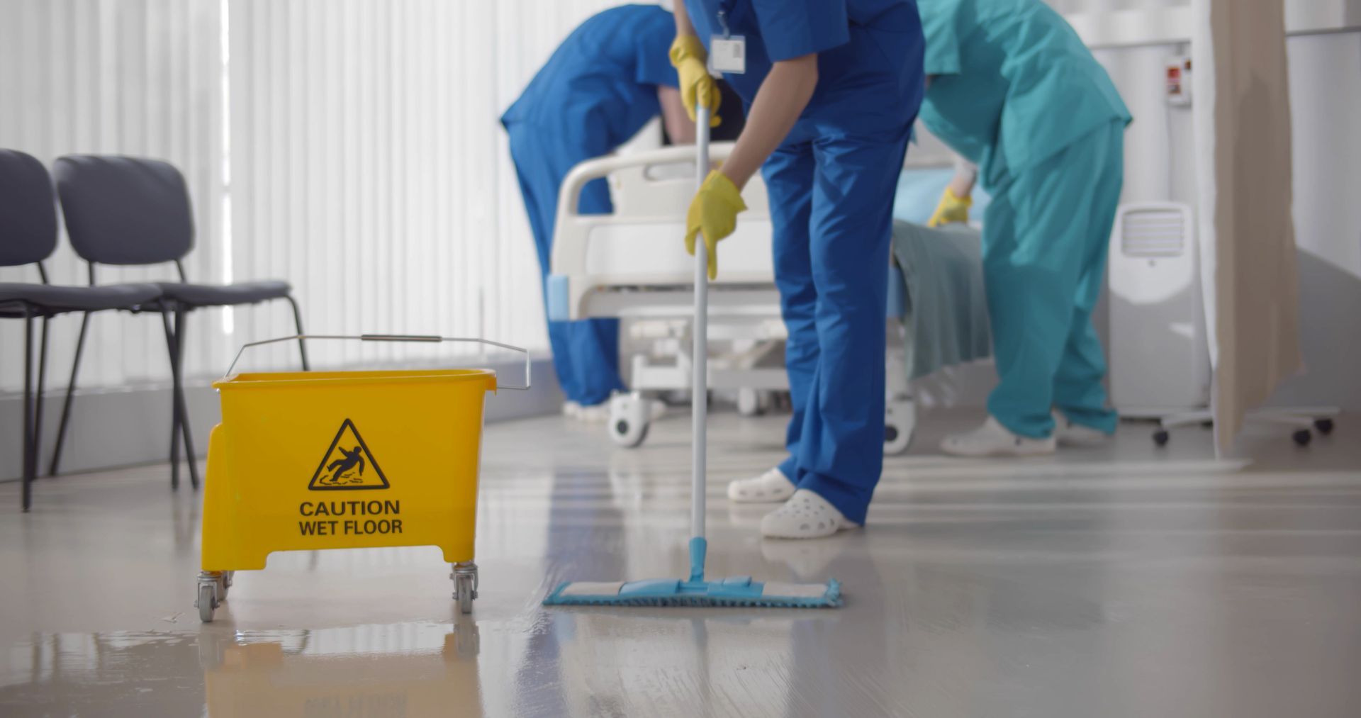 Janitorial staff mopping floors and making a bed in an empty hospital ward.