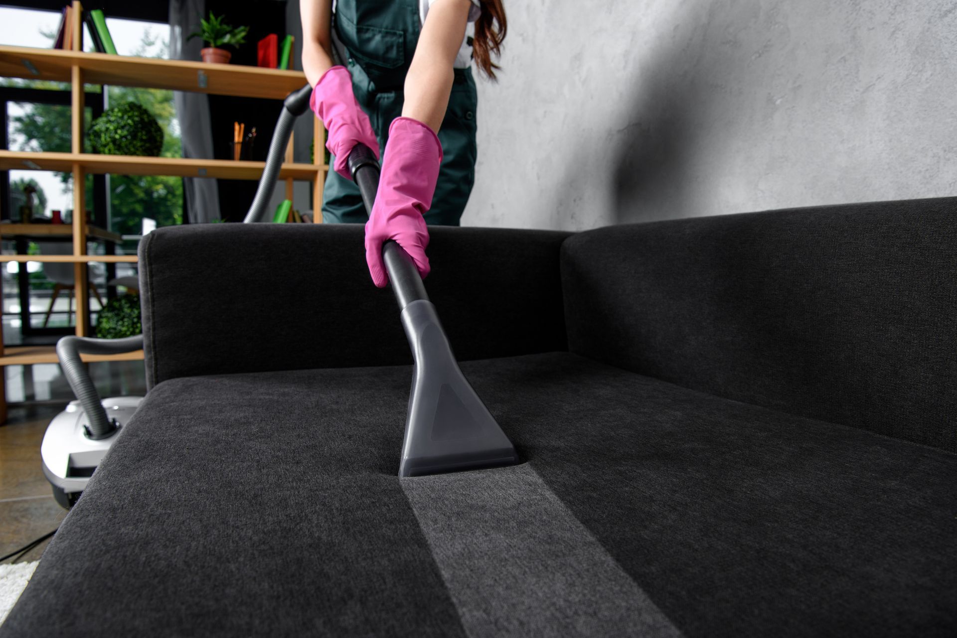 Person in pink gloves vacuums a black sofa, leaving a clean gray strip.