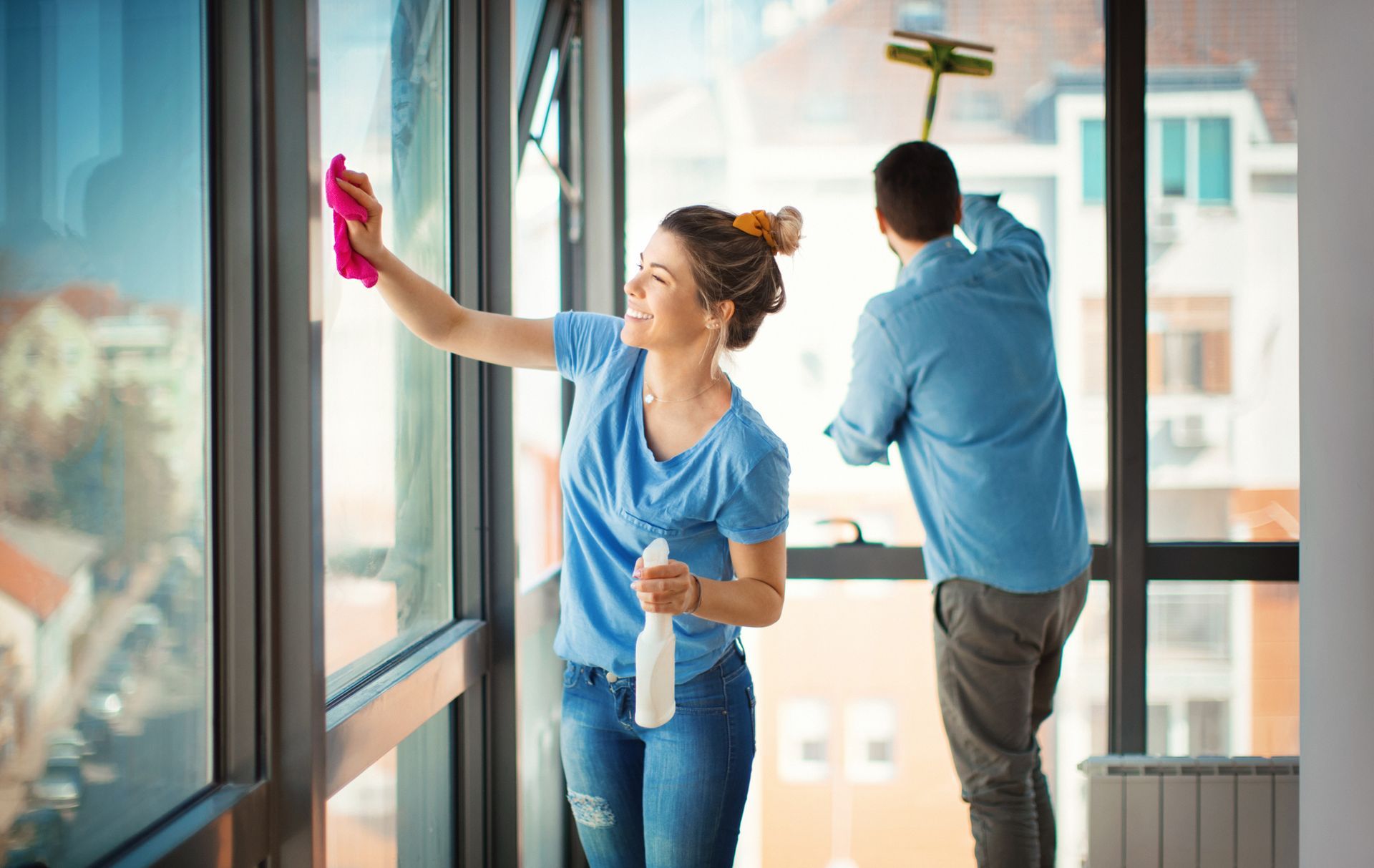 Woman spraying and wiping window while man cleans with squeegee in bright room.
