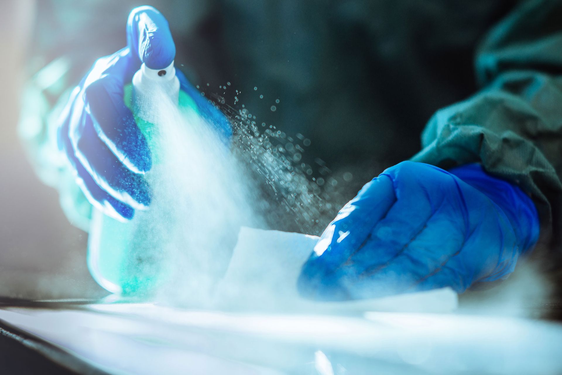 Hands in blue gloves spraying disinfectant onto a white wipe.