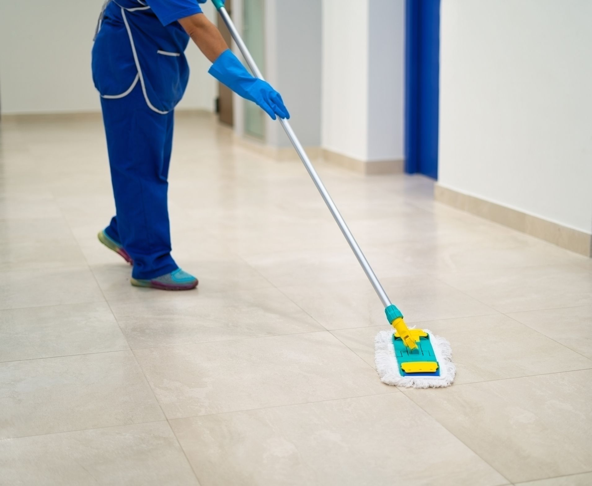 Person in blue scrubs mopping a beige tiled floor in a white hallway.