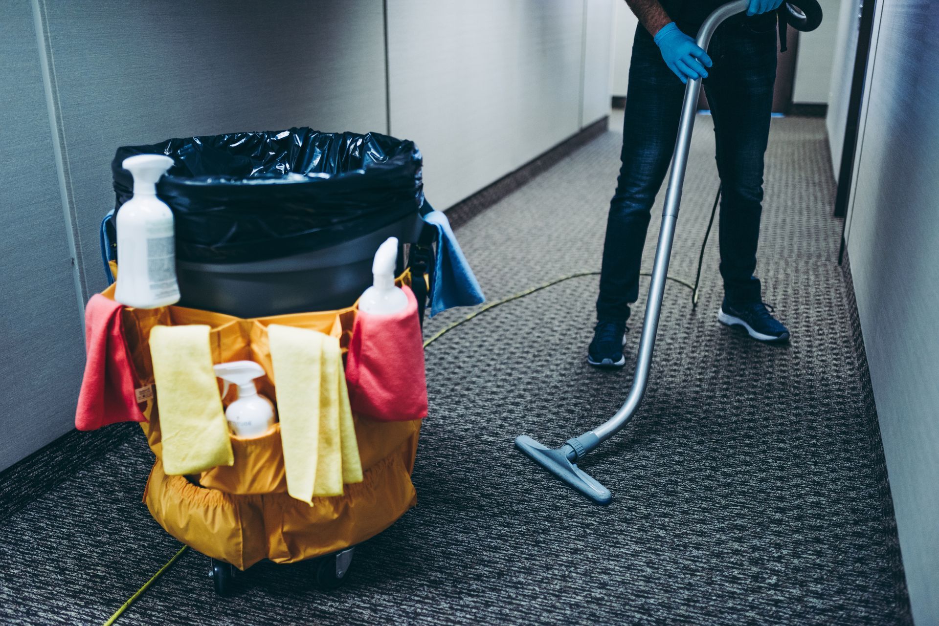 Person vacuuming carpeted hallway next to a cleaning cart with supplies and trash bag.