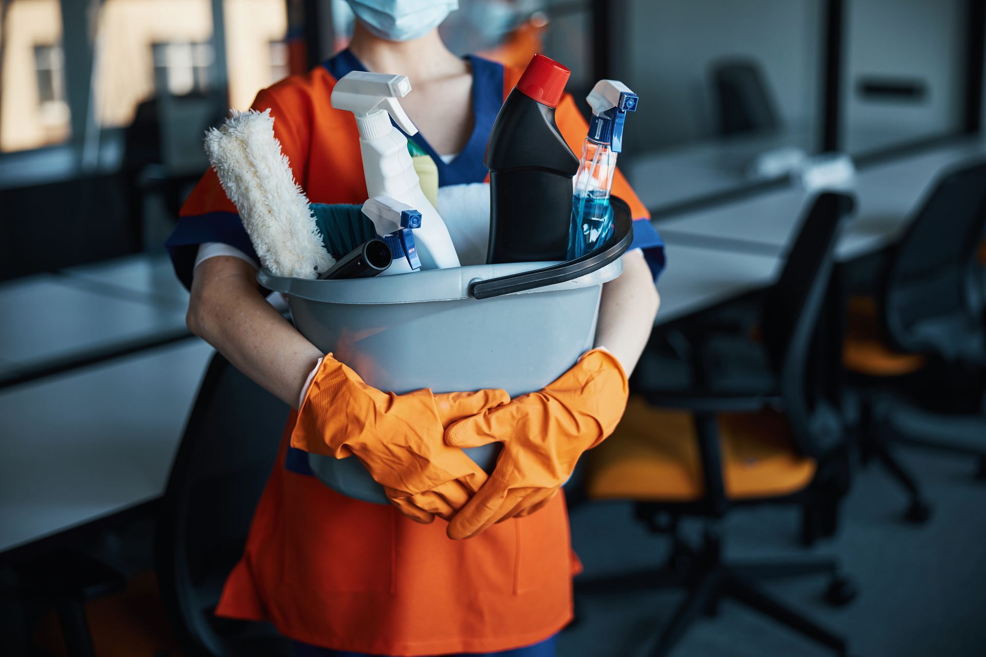Person in uniform wearing gloves and a mask holding a bucket of cleaning supplies in an office setting.