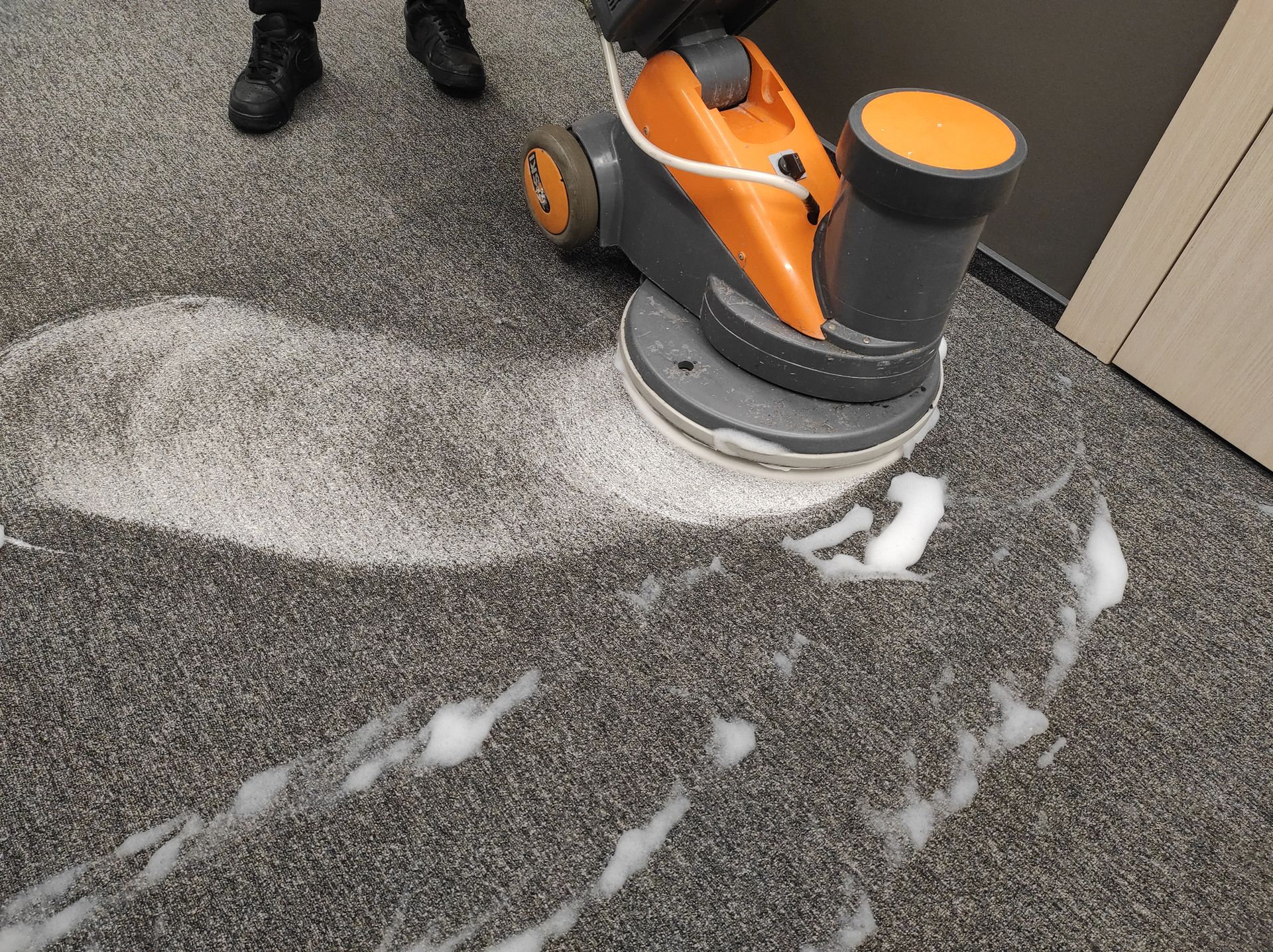 Person using an orange and gray floor cleaner on a gray carpet, creating white foam.