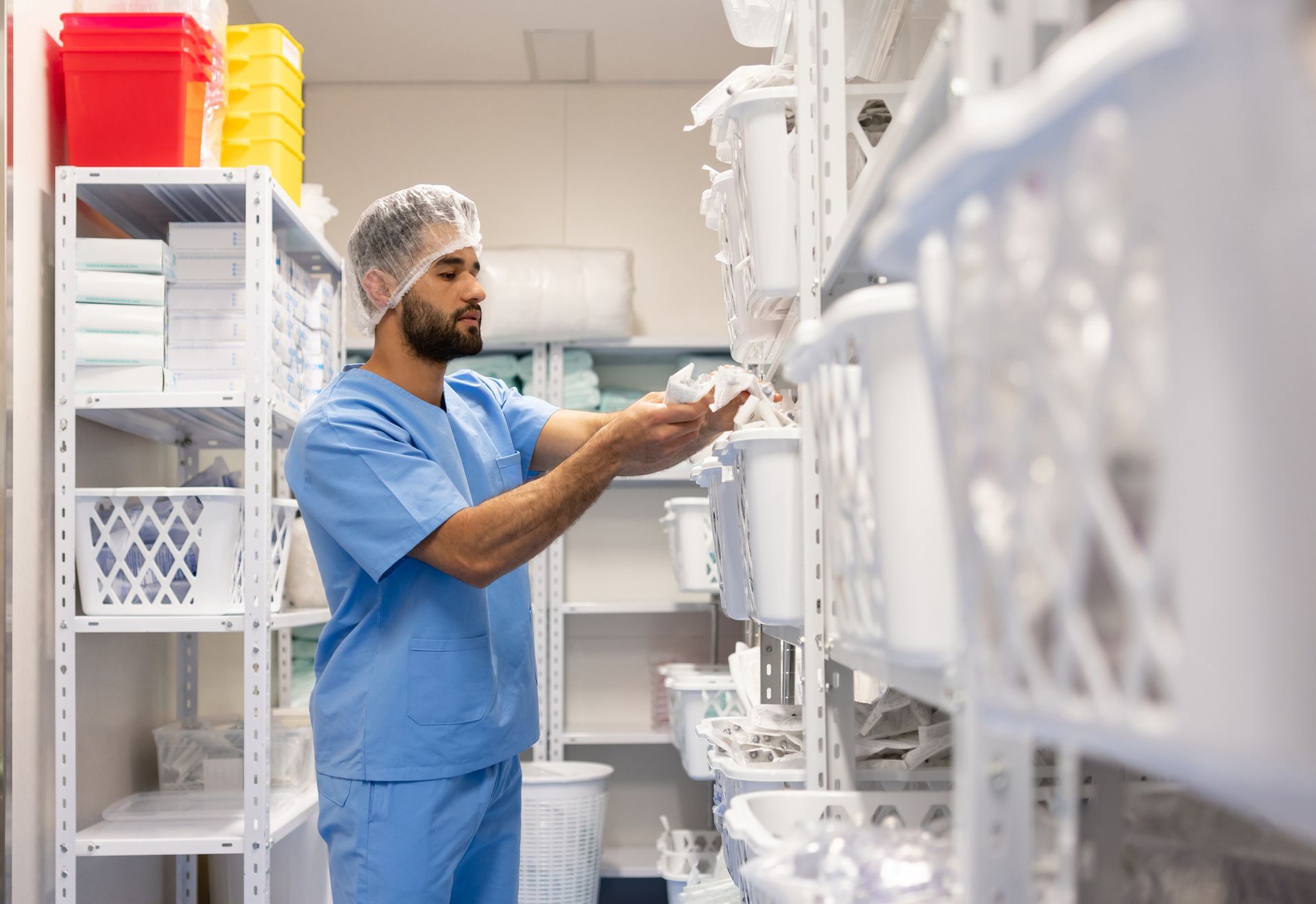 Man in scrubs, hair net stocking supplies in a medical storage room.