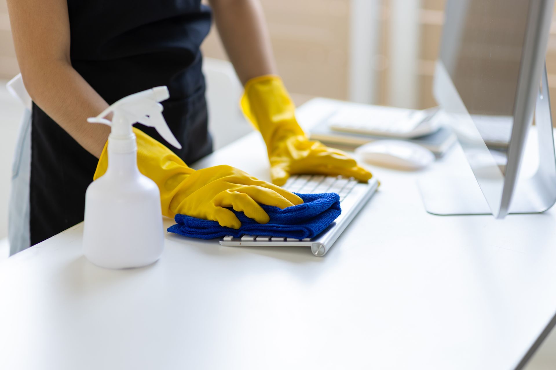 Person wearing yellow gloves cleaning a keyboard with a blue cloth and spray bottle on a white desk.