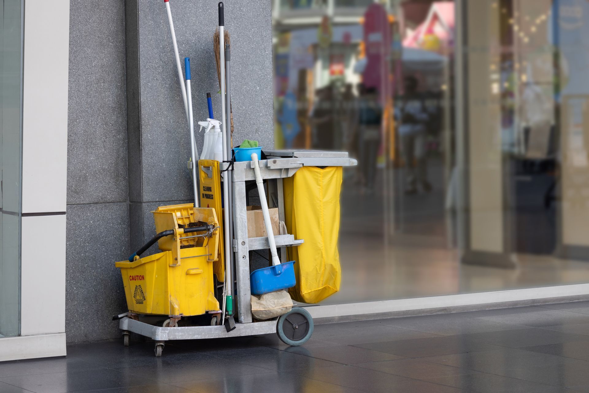 Cleaning cart with yellow buckets, mops, and brooms; resting beside a building's glass entrance.