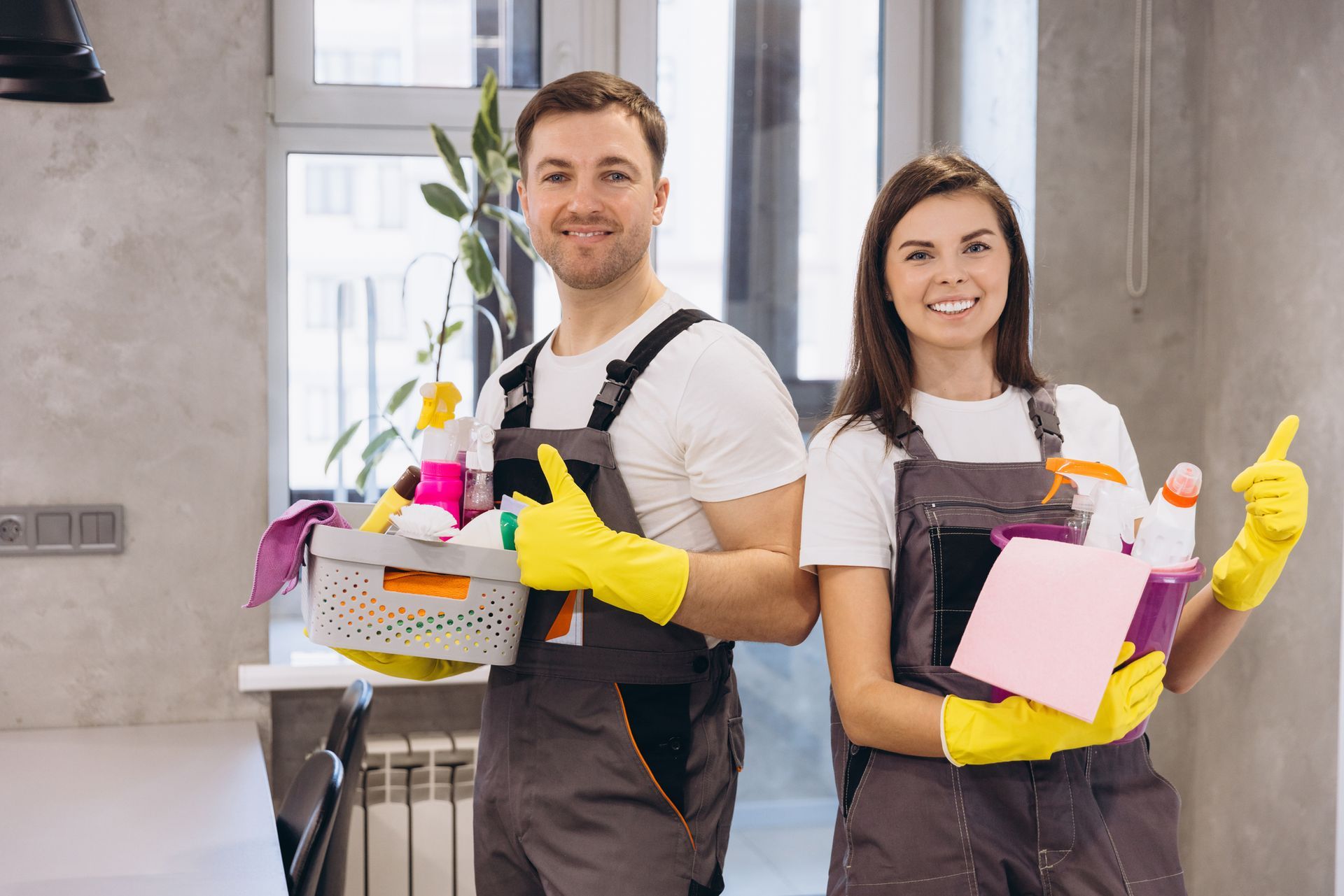 Two smiling cleaners in coveralls and gloves holding cleaning supplies.