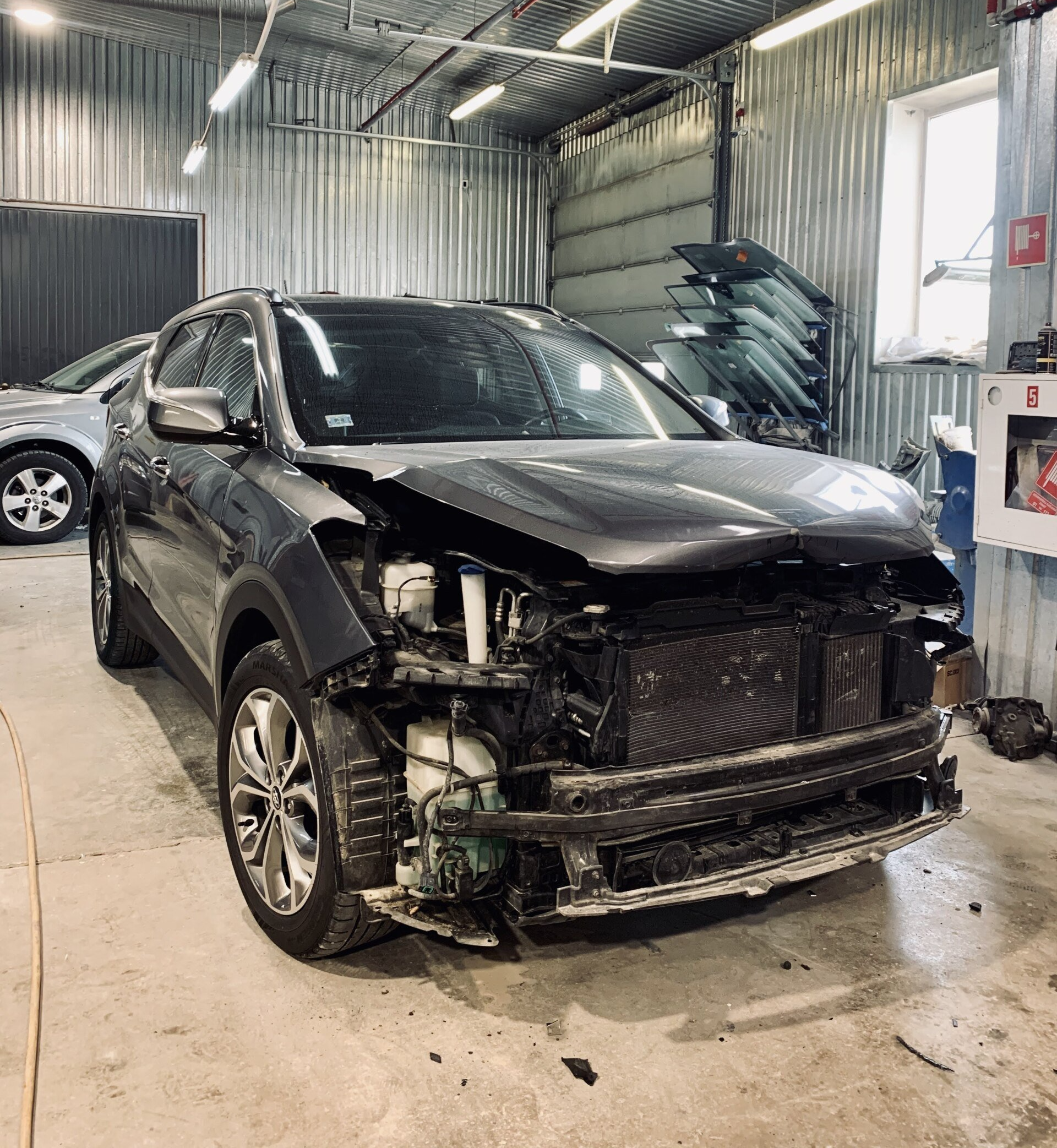 Bojāta automašīna ir novietota garāžā ar paceltu motora pārsegu.