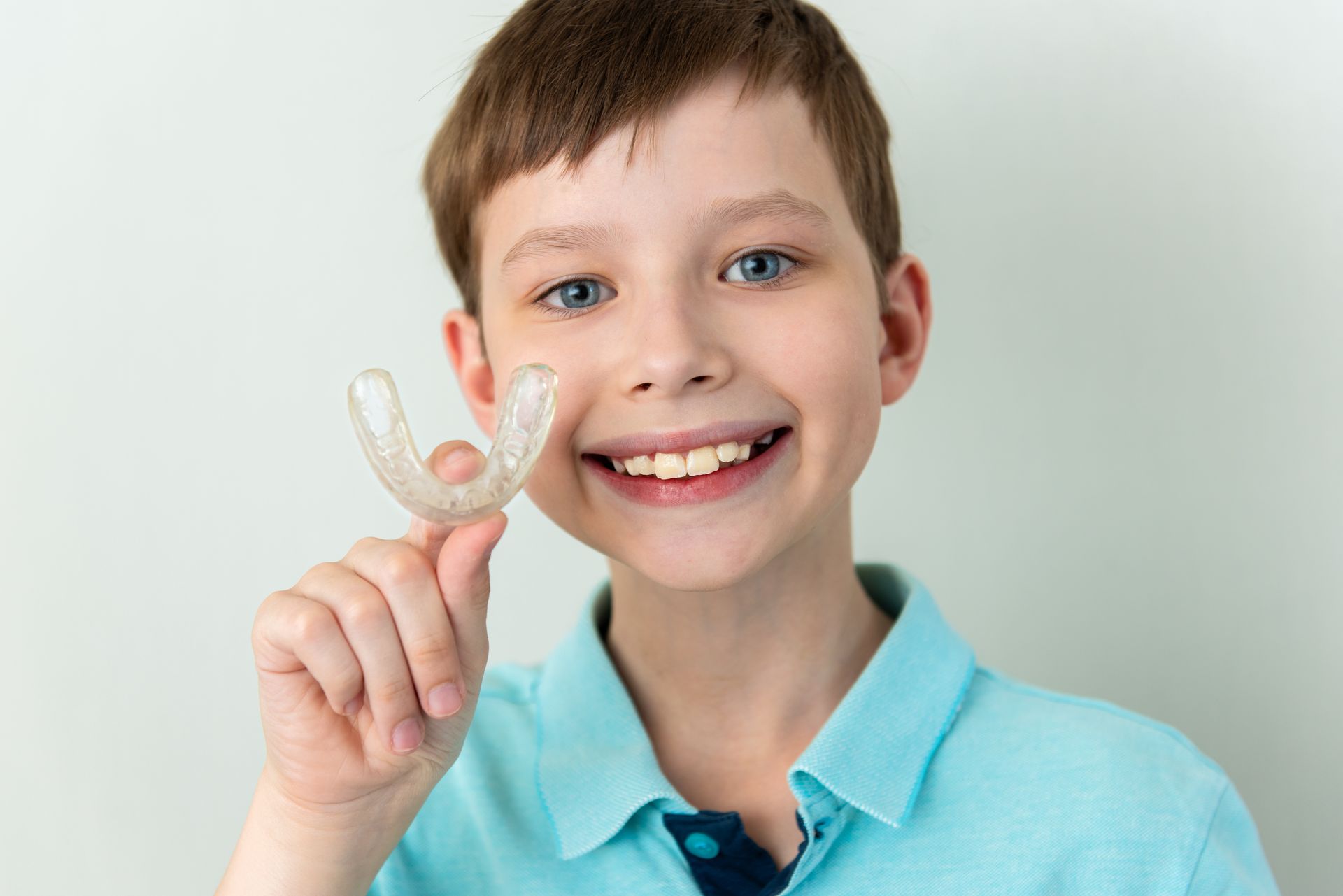 A smiling boy smiles as he holds an Invisalign retainer, standing in front of a white background.