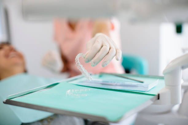 Gloved hand holding a clear dental aligner above a tray in a dental clinic setting.