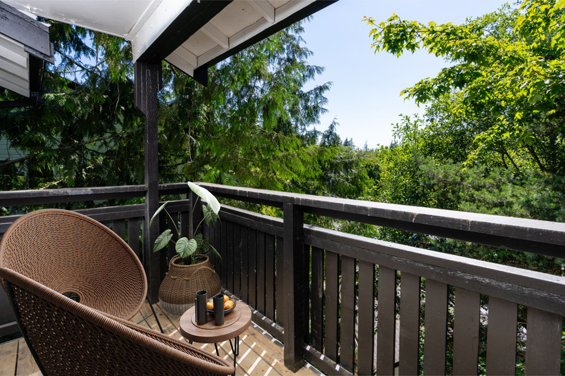 Balcony with wicker chair, potted plant, and small table, surrounded by lush green trees.