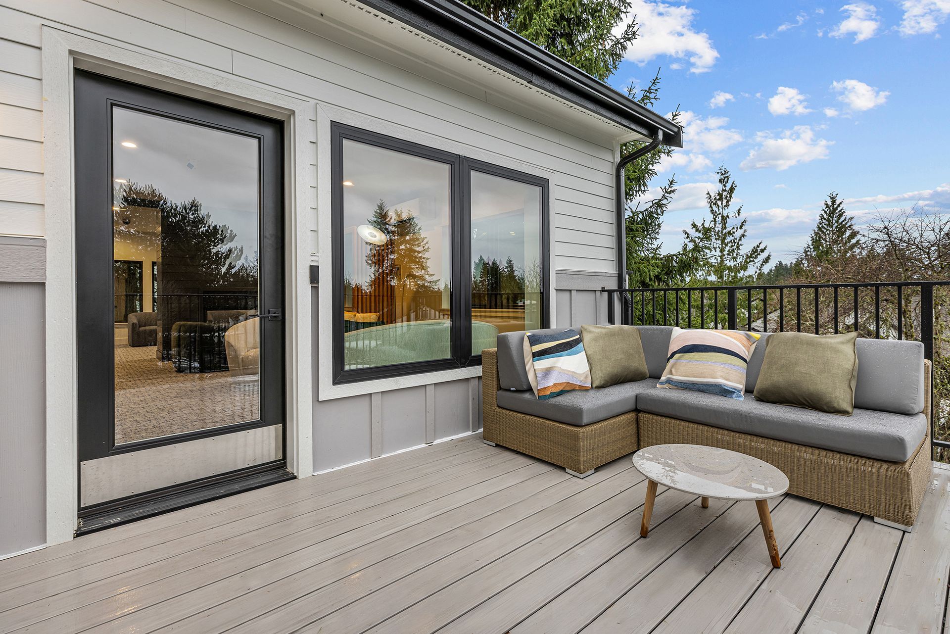 Gray house with a deck and wicker sofa overlooking trees; sunny day.
