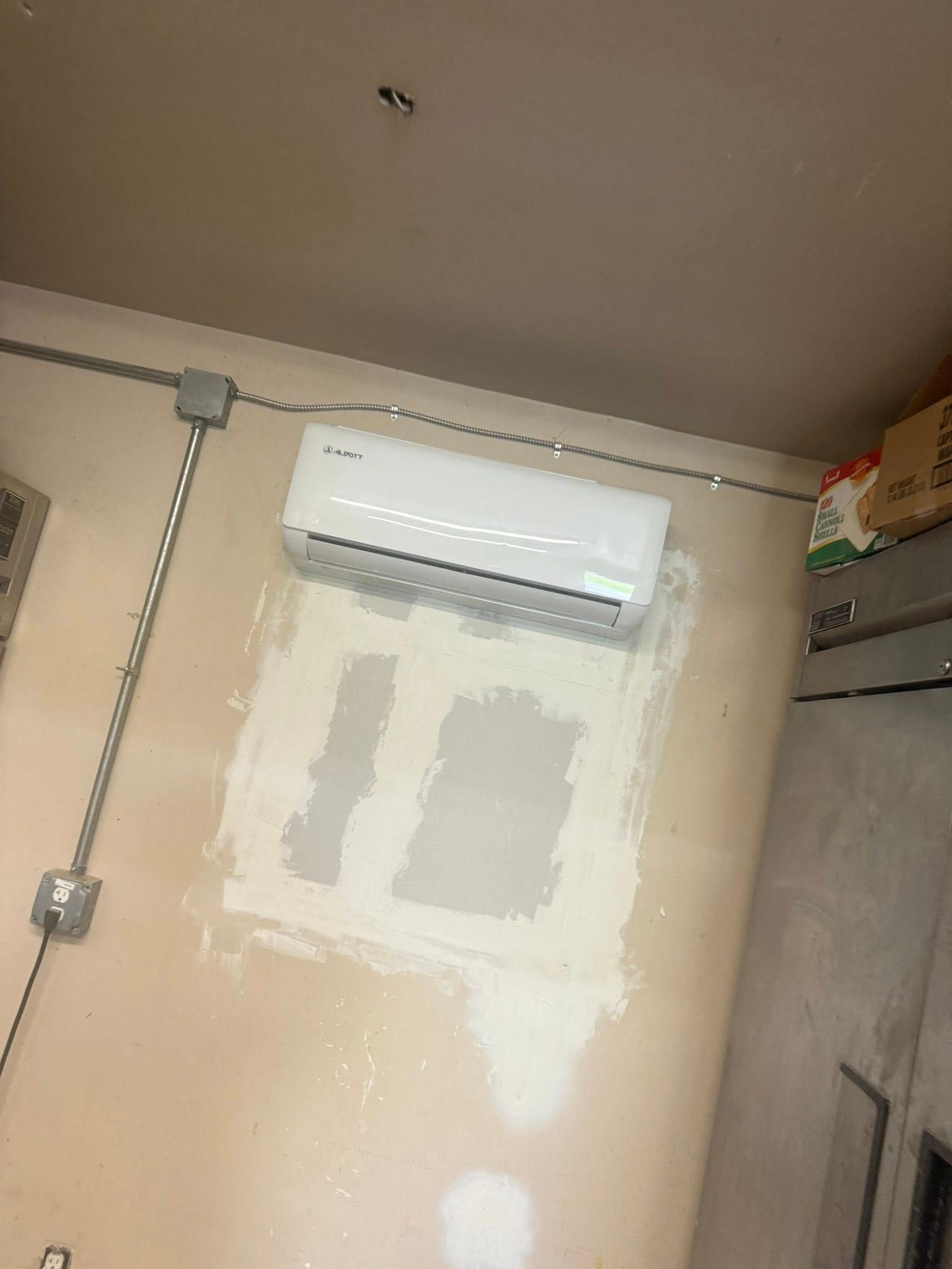 Air conditioner mounted on a wall with electrical conduit, surrounded by patched drywall.