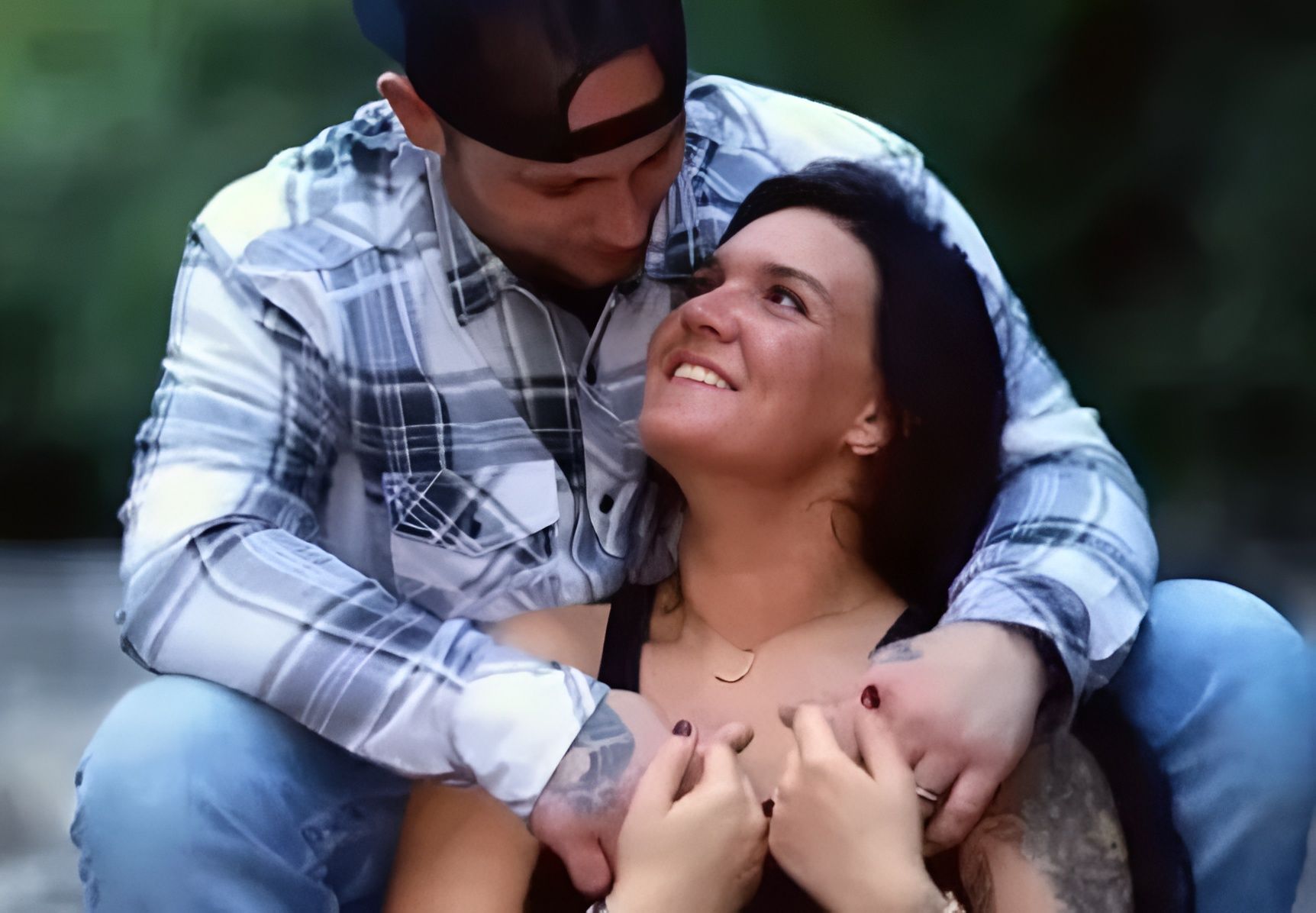Man hugging woman, both smiling. They're outdoors, man in plaid shirt, woman in black, appears to be daytime.