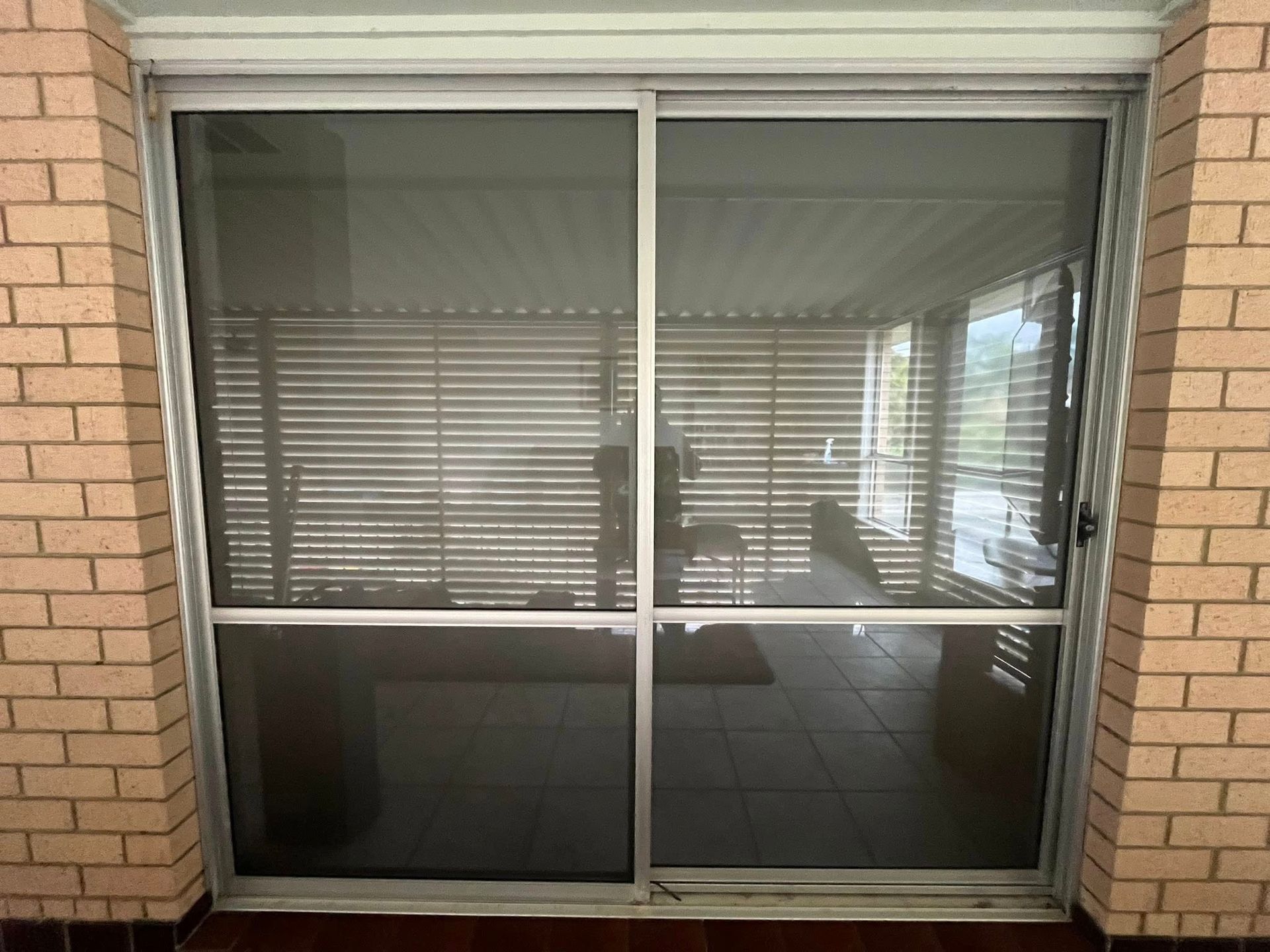 Sliding glass door with screen set in brick, reflecting a room with blinds and outdoor view.