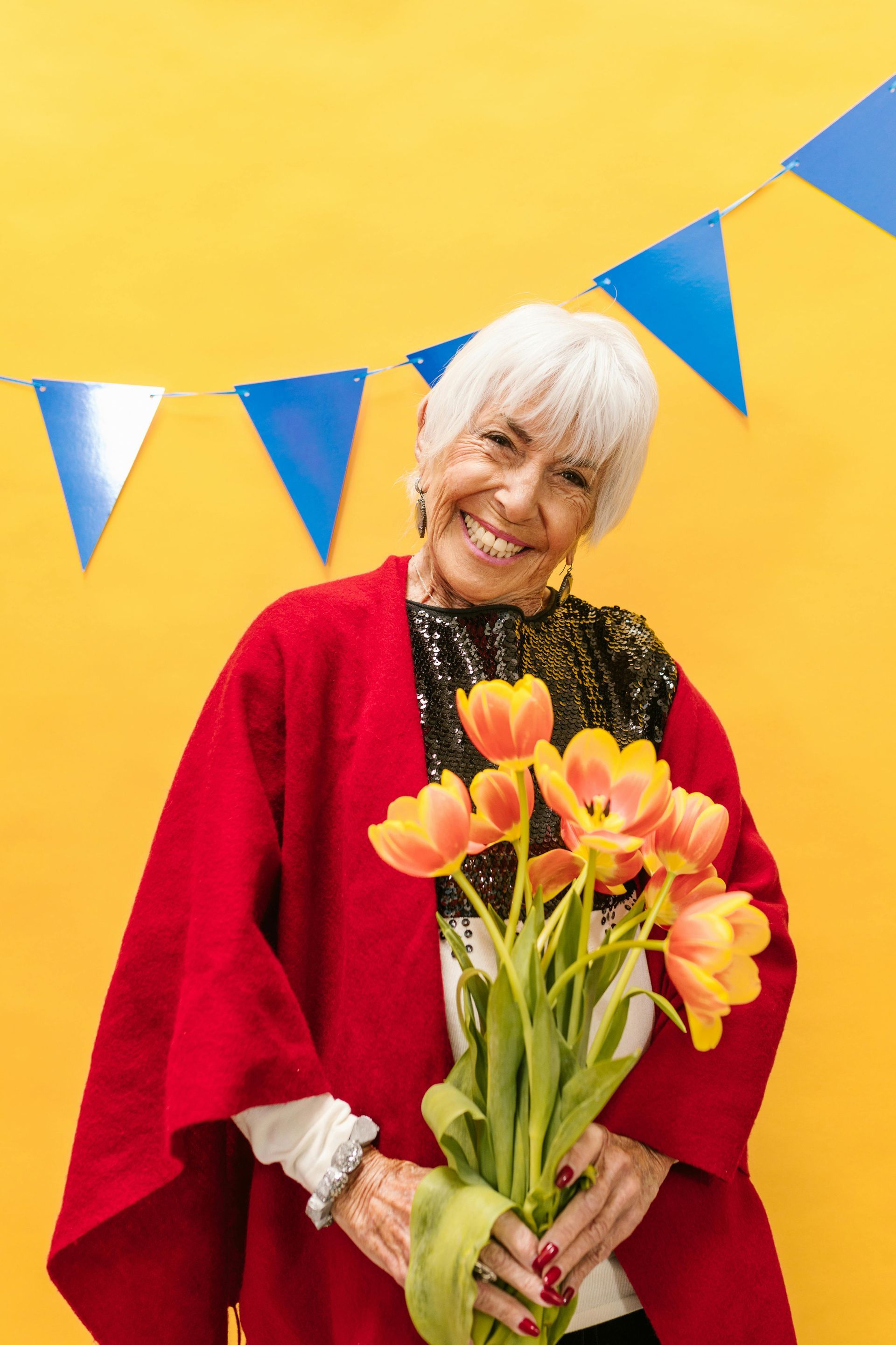 An elderly woman is holding a bouquet of flowers in front of a yellow wall.