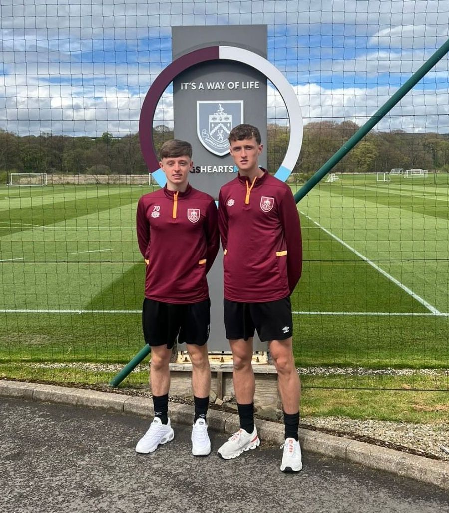 Two young men are standing next to each other in front of a soccer field.
