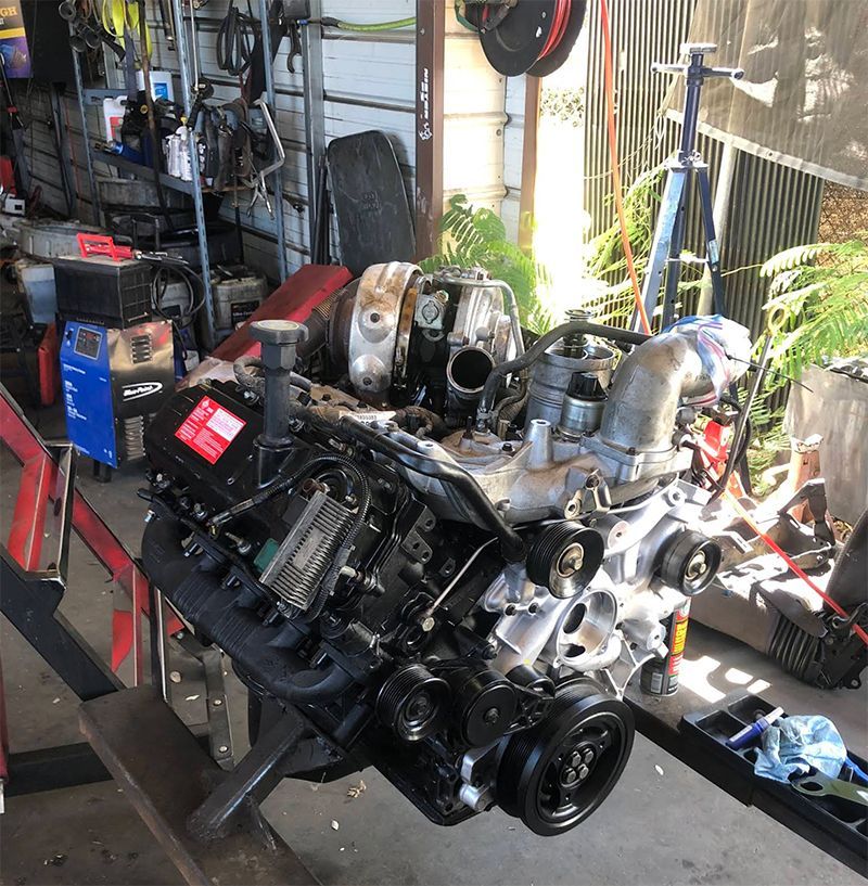 Diesel engine on a stand, in a garage. Black and silver components, red battery, tools visible.