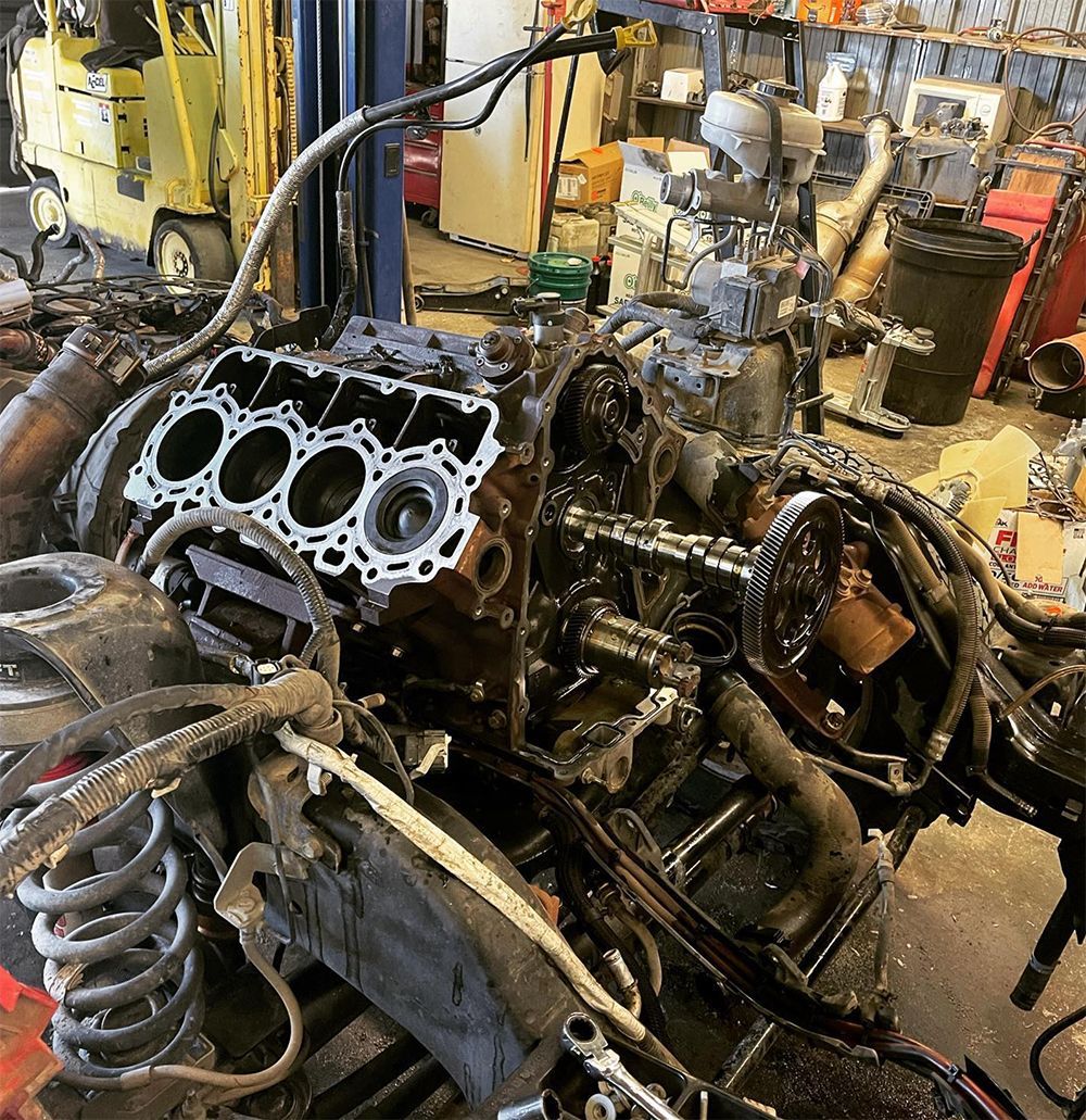 Engine parts disassembled in a cluttered mechanic's shop. Cylinders, wiring, and tools are visible.