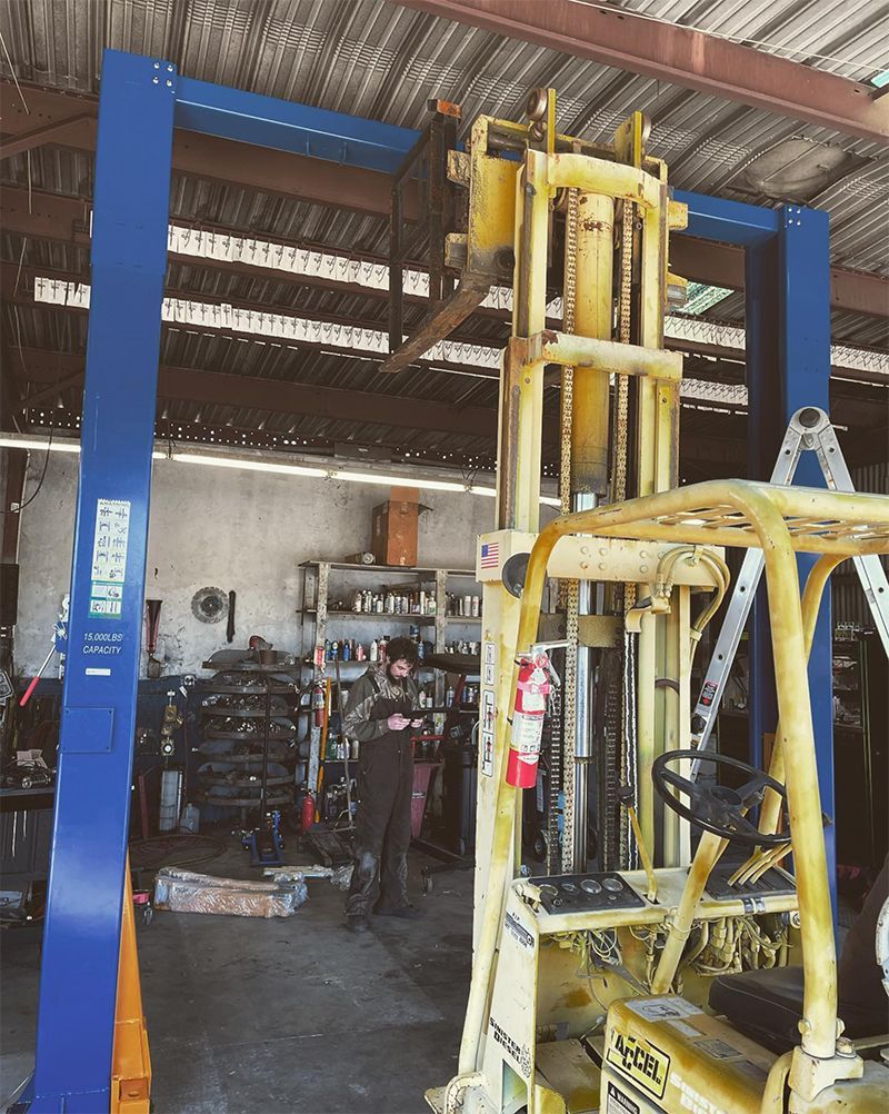 A person stands between a blue car lift and yellow forklift in a garage.