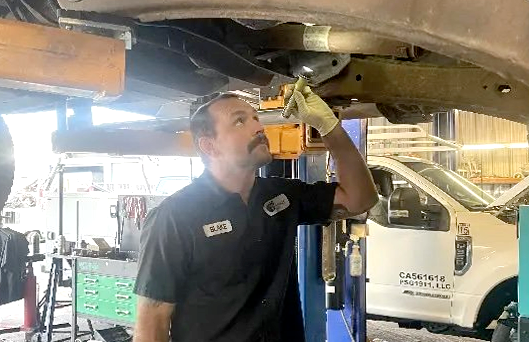 Mechanic inspecting underside of car raised on a lift in a garage. He wears a work shirt and looks upward.