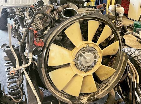 Truck engine with large fan, various wires, and hoses in a garage setting.