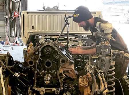 Mechanic working on a truck engine in a garage. He wears a beanie and focuses on the engine components.